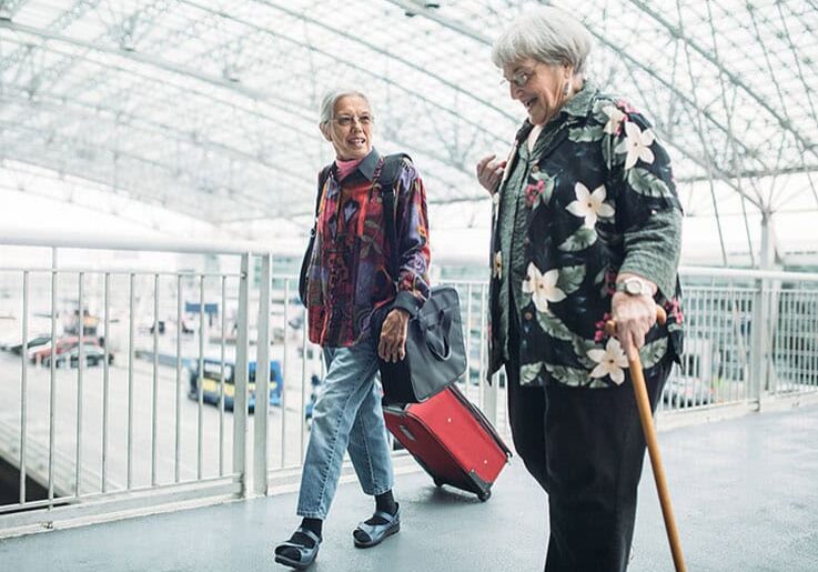Two mature friends in their 70's walk through the airport with luggage in tow smiling as they talk about their trip.  One woman uses a walking cane for assistance.